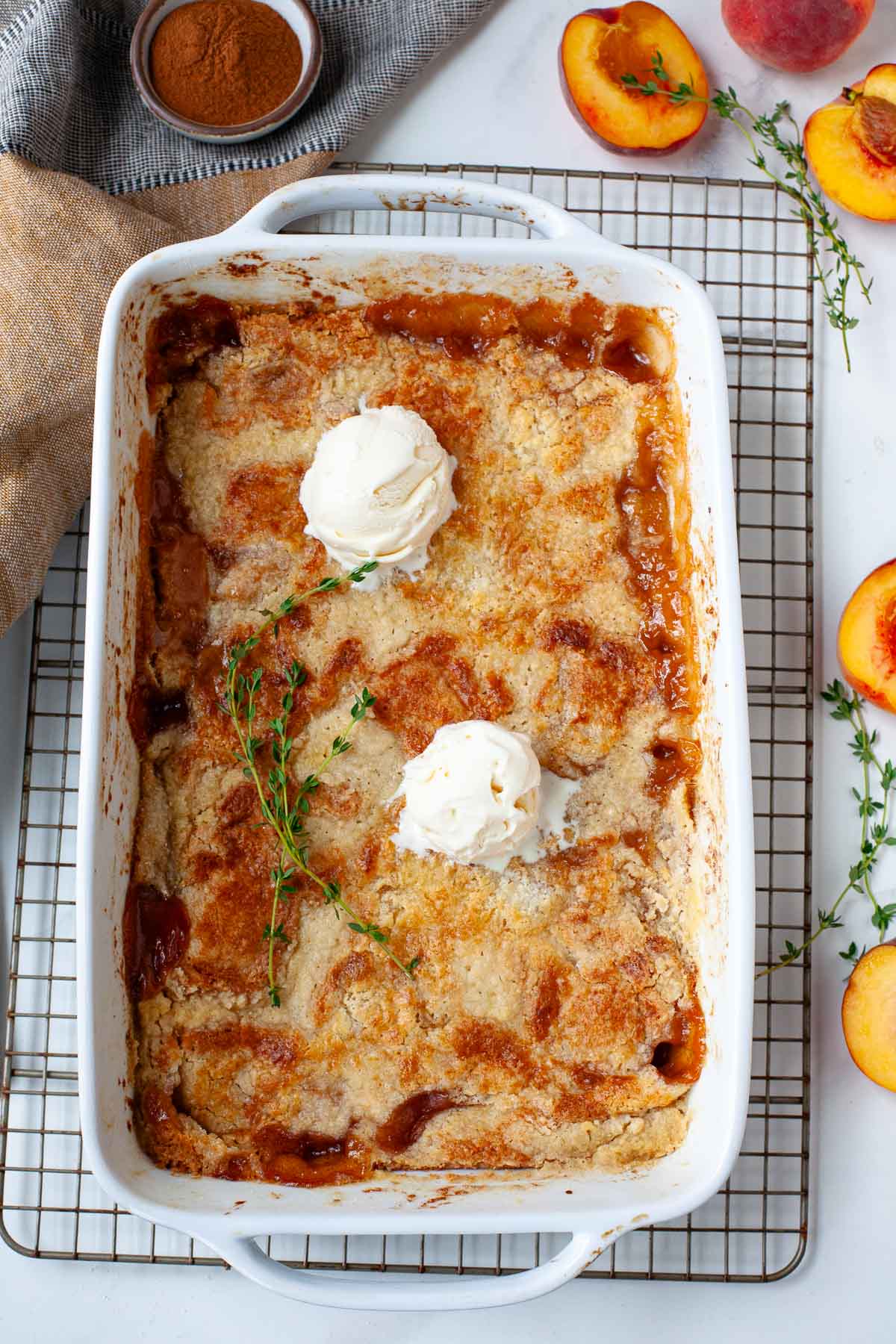 cake mix peach cobbler in white baking dish on wire rack topped with two scoops of vanilla ice cream and fresh thyme sprigs