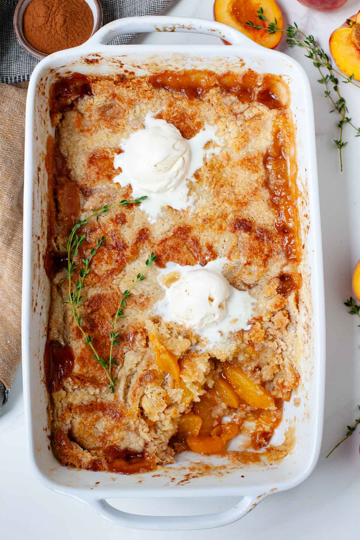 cake mix peach cobbler in white baking dish topped with two scoops of vanilla ice cream garnished with sprigs of thyme